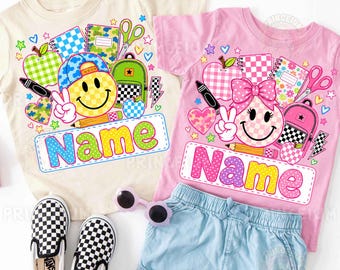 May include: Two short-sleeve t-shirts, one cream and one pink, featuring colorful school-themed graphics including smiley faces, backpacks, scissors, and notebooks. Each shirt displays the word Name in a large, multi-colored bubble font.