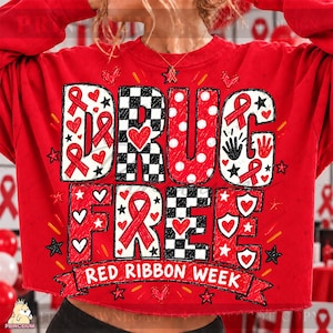 Pode incluir: Uma camisola vermelha de manga comprida com o texto DRUG FREE e RED RIBBON WEEK. As letras estão decoradas com fitas vermelhas, padrões de xadrez preto e branco, bolas brancas, corações, estrelas e gráficos de impressões de mãos.