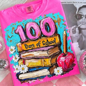 May include: A bright pink t-shirt with a colorful graphic celebrating 100 days of school. The design features the number "100" in pink, stacked books, a red apple, a pencil, and the text "Days of School".