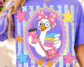 May include: A purple t-shirt featuring a graphic of a white goose wearing a pink floral bow and round glasses. The goose holds a pink polka-dot cup and a stack of colorful books while wearing a pink satchel. The design includes stars and stripes.