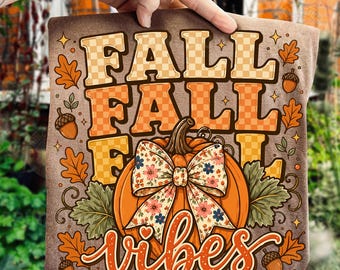 May include: A brown t-shirt featuring the text FALL FALL FALL in a checkered orange pattern, accompanied by a large orange pumpkin with a floral patterned bow, surrounded by illustrations of autumn leaves and acorns with the word vibes below.