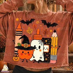 Op de afbeelding: Een roestkleurig sweatshirt met lange mouwen en een grafische print van een kleurpotlood, een pompoen met een heksenhoed, een spook met een spinnenweb, een zwart notitieboek en een potlood, omringd door zwarte vleermuizen, oranje sterren en herfstbladeren.
