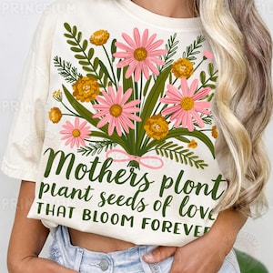 May include: Cream-coloured t-shirt with a floral bouquet design. The bouquet features pink and yellow flowers, green leaves, and the text "Mothers plant seeds of love THAT BLOOM FOREVER" in green script, tied with a pink ribbon.