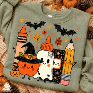 Op de afbeelding: Een olijfgroene sweater met een grafische print van een lachende oranje pompoen met een heksenhoed, een wit spookje, een geel potlood en een oranje waskrijtje, aangevuld met zwarte vleermuizen, herfstbladeren en kleine sterretjes.