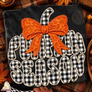 Può includere: Una maglietta grigio scuro con una grande grafica di zucca a quadri bianchi e neri. Un fiocco in pizzo arancione è fissato sulla parte superiore della zucca. Il testo PUMPKIN SEASON è scritto in lettere a quadri bianchi e neri.