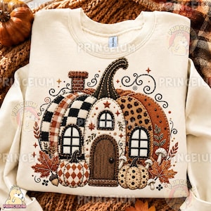 Op de afbeelding: Een crèmekleurig sweatshirt met ronde hals en een grote, gedetailleerde afbeelding van een pompoenvormig huis. Het ontwerp bevat een bakstenen schoorsteen, een houten deur, ramen en kleinere pompoenen aan de basis met ruitpatronen.