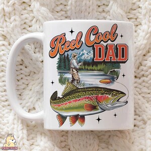 May include: White ceramic mug with a fishing theme. The mug features the text "Reel Cool Dad" in orange and red, along with an illustration of a fisherman and a large trout. The design includes a mountain and lake scene.