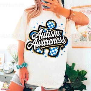 May include: White t-shirt featuring a blue and white chequered puzzle piece design with the text "Autism Awareness" in gold glitter lettering.