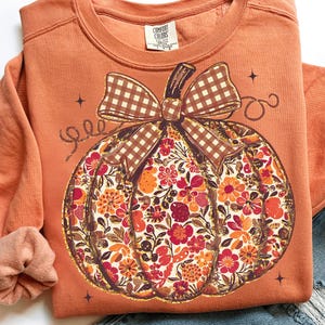 May include: Orange sweatshirt featuring a floral pumpkin design with a brown gingham bow. The pumpkin is filled with orange, red, and yellow flowers. The sweatshirt has a crew neck and long sleeves. The Comfort Colors brand tag is visible.
