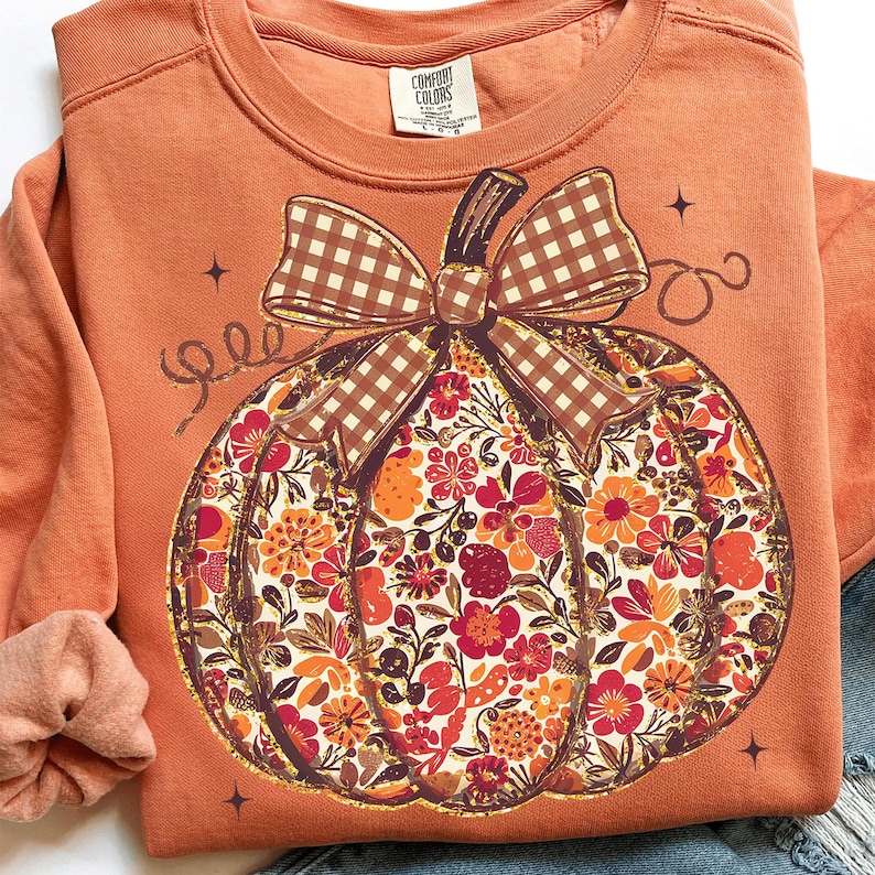 May include: Orange sweatshirt featuring a floral pumpkin design with a brown gingham bow. The pumpkin is filled with orange, red, and yellow flowers. The sweatshirt has a crew neck and long sleeves. The Comfort Colors brand tag is visible.