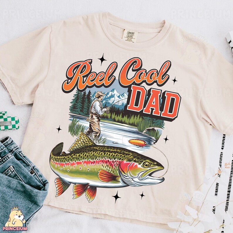 May include: Cream-colored t-shirt featuring a graphic of a fisherman, a trout, and the words "Reel Cool Dad" in orange and red. The design includes a mountain and river scene, with black star accents.