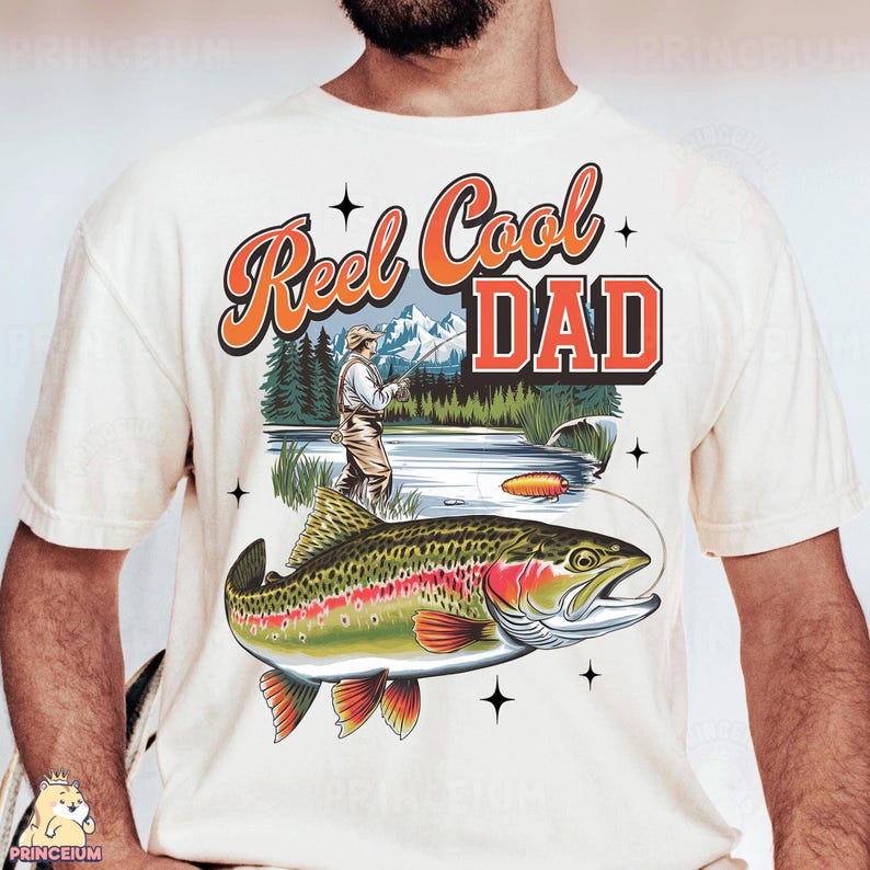 May include: Cream-colored t-shirt featuring a colorful graphic of a trout and a fisherman. The text "Reel Cool DAD" is in orange and red. The design includes a lake, mountains, and trees, with a fishing lure in the water.