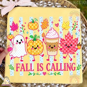 May include: A light yellow t-shirt featuring a graphic print of a ghost, a checkered pumpkin, a coffee cup with whipped cream, and a checkered maple leaf. The text "FALL IS CALLING" is printed in pink below the characters, surrounded by small hearts.