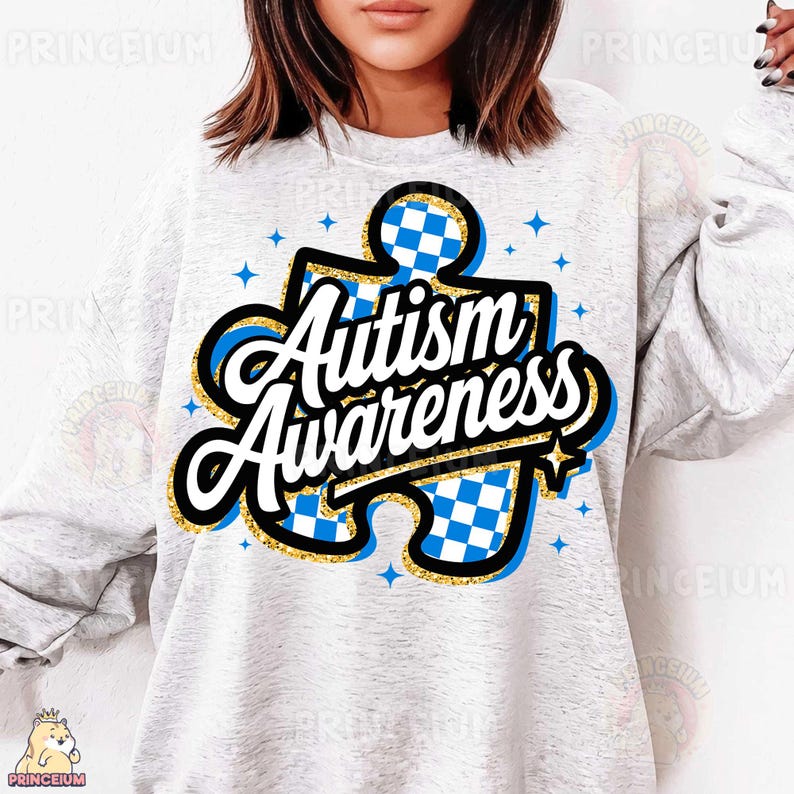 May include: A grey sweatshirt featuring a blue and white chequered jigsaw piece design. The text "Autism Awareness" is written in gold glitter lettering on the jigsaw piece.