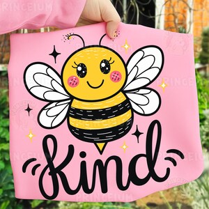 May include: Pink fabric with a cartoon bee illustration. The bee is yellow and black with white wings, rosy cheeks, and a smiling face. The word "Kind" is written in black script below the bee, with star accents.