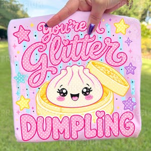May include: A light pink t-shirt featuring a cute cartoon dumpling with a smiling face inside a bamboo steamer. The text "You're a Glitter Dumpling" is written in pink with white polka dots, surrounded by stars.