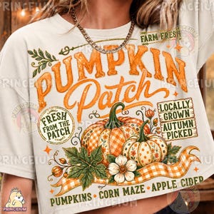 May include: A cream-colored t-shirt featuring a graphic print with the text "Pumpkin Patch," "Fresh From The Patch," "Locally Grown," "Autumn Picked," "Pumpkins," "Corn Maze," and "Apple Cider" alongside illustrations of pumpkins and leaves.