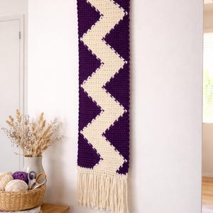 May include: A handmade, rectangular wall hanging with a chevron pattern. The design features a deep purple background with a cream-colored zigzag pattern. The bottom edge has a cream-colored fringe. The wall hanging is displayed on a white wall.