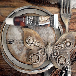 Könnte beinhalten: Eine Steampunk-Schmetterlingsskulptur mit filigranen Zahnrad-Details liegt auf einer rustikalen, runden Holzoberfläche. Eine Tube Sepia Antique Wax liegt auf der Oberfläche. Eine Gabel und ein Löffel sind im Hintergrund zu sehen.