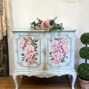 Könnte beinhalten: Ein hellblauer Schrank mit goldenen Akzenten und Blumendesigns. Der Schrank hat zwei Türen mit je einem Quasten. Der Schrank steht auf einem hellen Holzboden. Der Text auf dem Schrank lautet "Est un beau endroit - est un lieu rempli d'amour".
