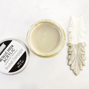 Puede incluir: Vista superior de una lata de pintura acrílica metalizada "Brushed Champagne", junto a un tarro abierto del mismo color y un elemento decorativo. La pintura es de un tono beige claro y brillante. La pieza decorativa está parcialmente pintada.