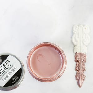 Könnte beinhalten: Eine Dose Metallic Sheen Acrylfarbe in Blush Chrome, neben einem offenen Behälter der gleichen Farbe und einer dekorativen weißen und blushfarbenen Applikation. Auf der Farbdose steht "METALLIC SHEEN ACRYLIC Blush Chrome 100 ml (3.4 fl. oz.)".