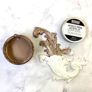 May include: An overhead shot features a metallic sheen acrylic paint in a silver tin, a decorative bronze-colored ornate scroll, and a round container filled with bronze paint. The text on the tin reads "Metallic Sheen Acrylic Polished Bronze".
