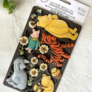 May include: A black silicone mold featuring characters from the "Olde Bear Lane" collection. The mold includes Winnie the Pooh, Piglet, Tigger, Eeyore, sunflowers, bees, and a honey pot. The packaging reads "Decor Moulds".
