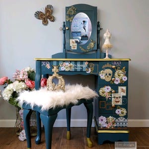 Op de afbeelding: Een teal kaptafel met bijpassende kruk, versierd met bloemen- en schedelstickers. De kaptafel heeft een spiegel, lades en gouden accenten. Een zacht wit kussen ligt op de kruk. Decoratieve vlinders zijn op de muur.