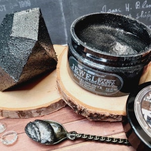 May include: A black jar of sparkling onyx jewel effect paste with a silver lid. The jar is sitting on a wooden coaster. The text on the jar reads "Jewel Effect Paste Sparkling Onyx 3.4 fl oz (100 ml)".