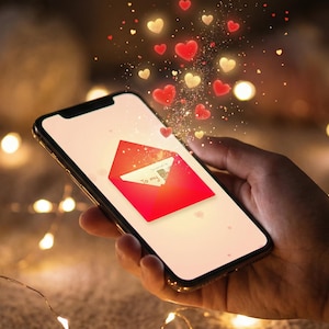 Puede incluir: Un teléfono inteligente muestra un sobre rojo con un mensaje, rodeado de formas de corazón rojas y doradas. El teléfono se sostiene en una mano, con un fondo borroso de luces, lo que sugiere un tema romántico.