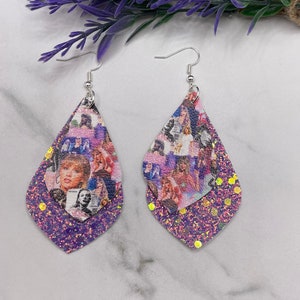 May include: A pair of dangle earrings with a purple glitter background and a layered image of a woman's face. The earrings are on a white marble surface.