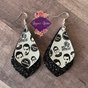 May include: A pair of dangle earrings with a black and white pattern. The top layer features a white background with black illustrations of faces and the text "EW, DAVID" and "ROSE APOTHECARY". The bottom layer is black glitter.