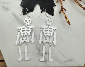 May include: A pair of white skeleton earrings with silver hooks. The earrings are dangling and feature a skeleton design with a skull at the top.