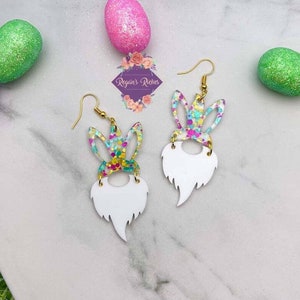 May include: A pair of white dangle earrings with a white beard and colorful bunny ears. The bunny ears are covered in glitter and have a white outline. The earrings are on a white background with glitter Easter eggs.