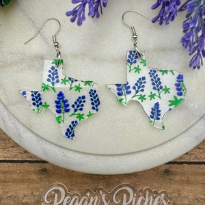 May include: A pair of clear acrylic earrings shaped like the state of Texas, with a bluebonnet floral pattern. The earrings have silver hooks.