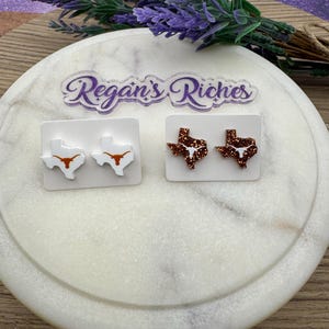 May include: Two pairs of Texas-shaped earrings. One pair is white with a burnt orange longhorn design, and the other is a glittery copper color with a white longhorn design. The earrings are displayed on white cards.