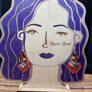 May include: A pair of dangle earrings with a Halloween theme. The earrings are made of red glitter and feature a collage of Halloween images. The earrings are hanging from a wooden mannequin head with purple hair.