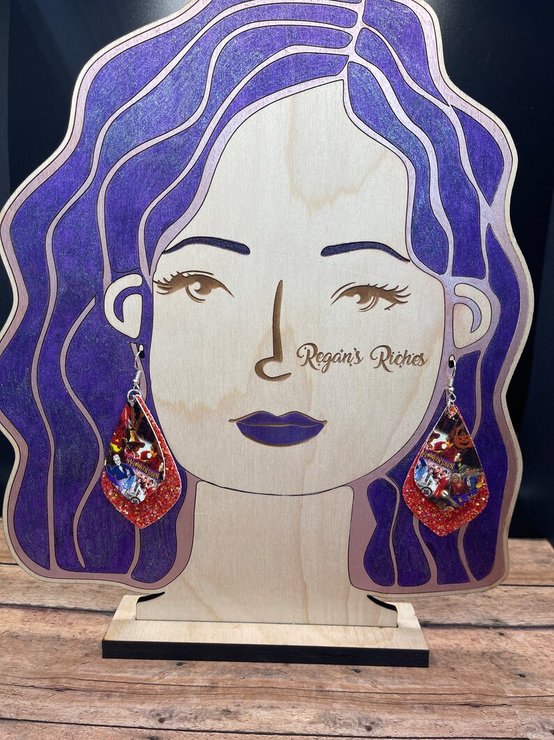 May include: A pair of dangle earrings with a Halloween theme. The earrings are made of red glitter and feature a collage of Halloween images. The earrings are hanging from a wooden mannequin head with purple hair.
