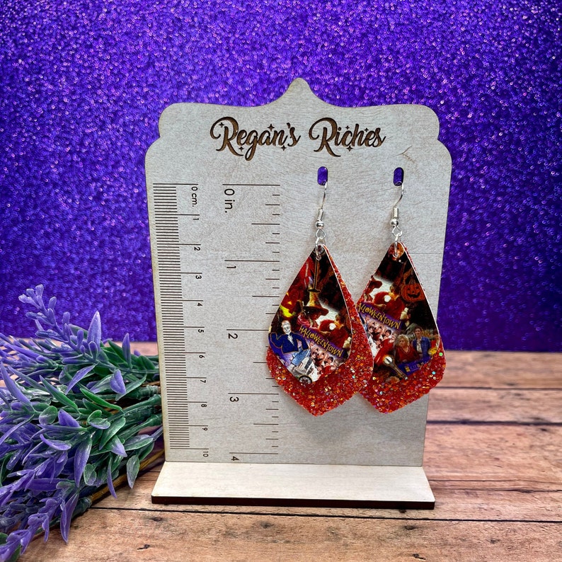 May include: A pair of red glitter teardrop earrings with a Halloweentown print. The earrings are on a wooden display stand with a ruler.