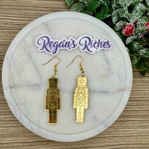 May include: Gold-colored dangle earrings featuring a nutcracker soldier design. Each earring has a hook for easy wear and a detailed depiction of the classic holiday figure. The earrings are displayed on a white marble surface with the text "Regan's Riches".
