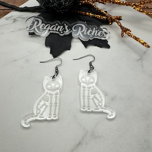 May include: A pair of clear acrylic earrings featuring a skeleton cat design. The earrings are silver in color and have a hook closure.
