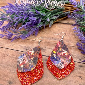 May include: A pair of red glitter dangle earrings with a double layer design. The top layer features a printed image of a scene from the movie "Hocus Pocus" with the text "Hocus Pocus" and "Salem".