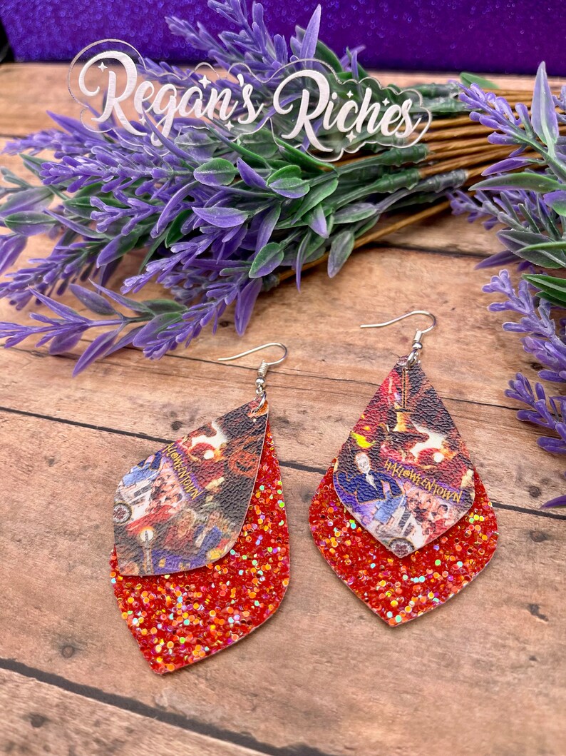 May include: A pair of red glitter dangle earrings with a double layer design. The top layer features a printed image of a scene from the movie "Hocus Pocus" with the text "Hocus Pocus" and "Salem".