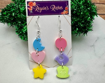 May include: A pair of dangle earrings with a pastel color scheme. The earrings feature a crescent moon, heart, star, and a hat with a flower on the top. The earrings are on a white card with the text "Regan's Riches".