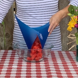 May include: A blue silicone watermelon corer and slicer being used to transfer watermelon chunks into a glass bowl. The bowl is on a red and white checkered tablecloth.