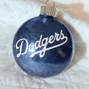 May include: A navy blue glitter Christmas ornament with the word "Dodgers" in white script. The ornament has a silver metal cap and a loop for hanging. The background is a white, textured surface.