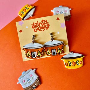 May include: A pair of dangle earrings and several decorative pins featuring a slow cooker design. The earrings and pins are in shades of orange, yellow, white, and brown. The earrings are on a yellow card with the text "dainty c me".
