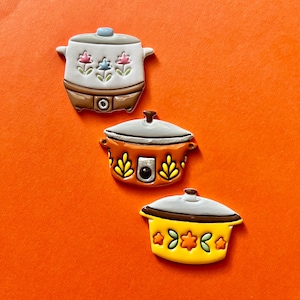 May include: Three colorful slow cooker-themed decorative items. The top one is white with pink and blue floral designs, the middle one is orange with yellow leaf designs, and the bottom one is yellow with green floral designs.