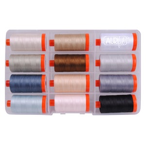 May include: A clear plastic storage case with 12 spools of thread. The spools are in various shades of gray, brown, beige, and black. The case has the word "Aurifil" printed on the top.